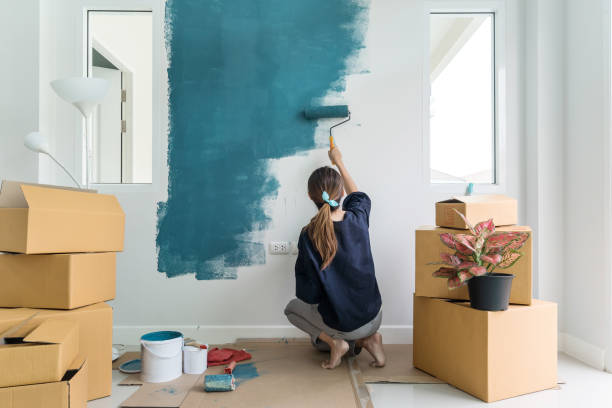 Человек красит стену в синий цвет в комнате с распакованными коробками и растением.