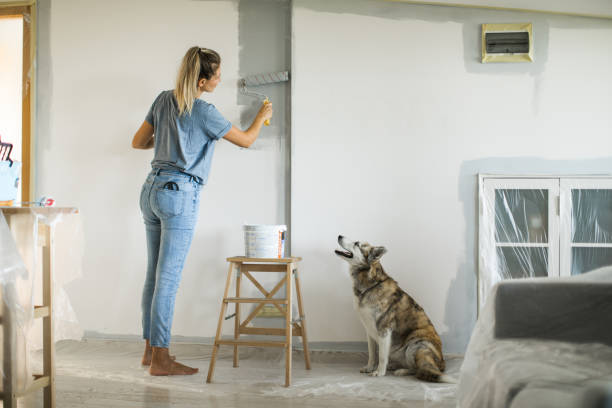 Женщина красит стену в комнате, а рядом сидит собака и наблюдает.