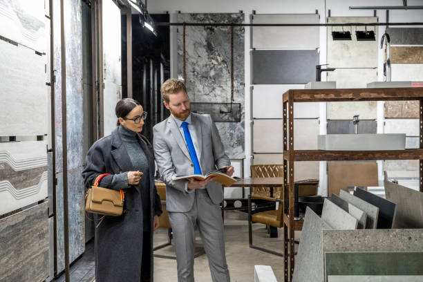 Мужчина в костюме и женщина в пальто рассматривают образцы настенной и напольной плитки в выставочном зале.