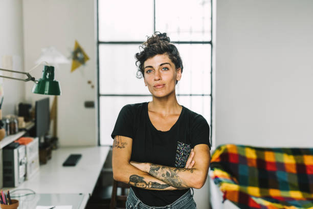 Женщина с татуировками уверенно стоит в домашнем офисе на фоне разноцветного одеяла.